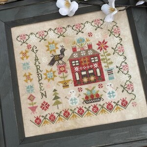 May include: A framed cross-stitch design featuring a red house with a white chimney and a blue roof. The house is surrounded by flowers and a bird. The text "GABBEN" is stitched in the upper left corner of the design. The frame is black and the design is on a cream-colored fabric.