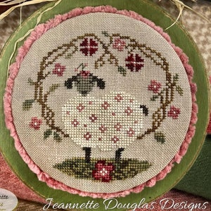 May include: A cross-stitch design of a white sheep with pink flowers on a green wooden plaque. The plaque has a pink and white fringe around the edge. The text "Chubby Sue" is written in cursive at the top of the plaque.