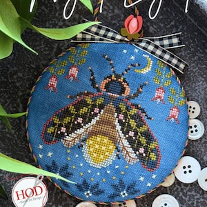 May include: A round, blue cross-stitch ornament featuring a detailed firefly design with a yellow abdomen and patterned wings. The ornament is adorned with a black and white checkered ribbon and the text "Night Light".