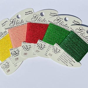 May include: A collection of colorful Glisten sparkling wool braid spools. The spools are in shades of yellow, peach, red, and two shades of green. Each spool is labeled with the brand name and product details.