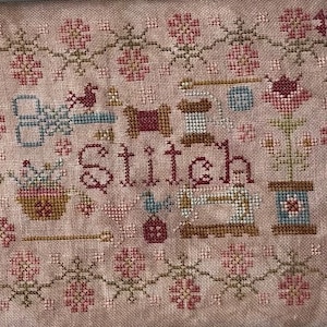 Peut inclure: Un sampler de point de croix avec une bordure florale et le mot "Stitch" au centre. Le sampler présente divers motifs sur le thème de la couture, notamment des ciseaux, des bobines de fil et une machine à coudre.