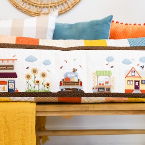 May include: A rectangular quilted lumbar pillow featuring a patchwork design with a central white panel depicting a fall-themed village scene. The scene includes a general store, sunflowers, a truck with pumpkins, a cider stand, and a house.