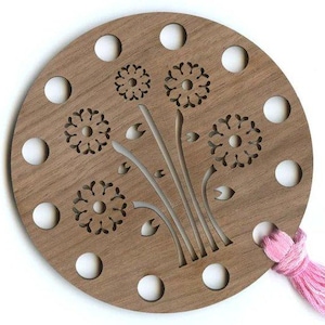 May include: A wooden round yarn bowl with a dandelion design and 12 holes around the edge. The bowl has a pink tassel attached to the side.