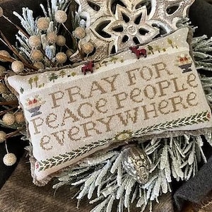 May include: A beige pillow with a cross-stitch design featuring the text "Pray for Peace People Everywhere". The pillow is surrounded by decorative elements, including a snowflake-shaped ornament, branches with small brown balls, and a snowy white plant.