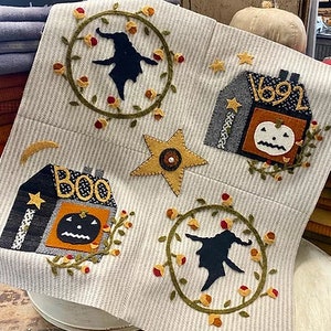 May include: A white fabric wall hanging with a Halloween theme. It features a witch, a house with the number 1692, a star, and a house with the word "BOO" on it. Each element is surrounded by a wreath of green leaves and red berries.