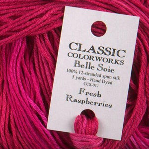 May include: A close-up of a skein of hand-dyed, fresh raspberry-coloured silk thread. A white tag reads "CLASSIC COLORWORKS Belle Soie" and lists the thread as 100% 12-stranded spun silk, 4.57 metres, CCS-011.