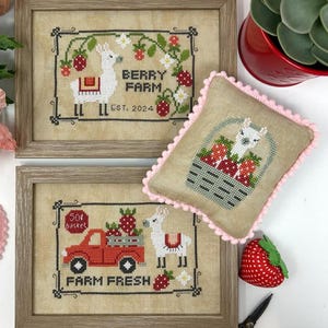May include: Two framed cross-stitch pieces and a small pillow with a llama and strawberry theme. One frame reads "Berry Farm Est. 2024". The other frame reads "Farm Fresh". The pillow has a pink pom-pom trim. A red strawberry and scissors are also visible.