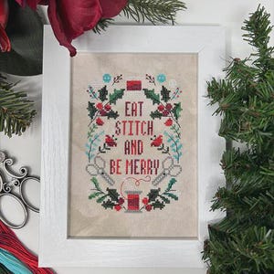 May include: A framed cross-stitch with the text "EAT STITCH AND BE MERRY" surrounded by a wreath of holly, berries, and scissors. The frame is white, and the background is a neutral beige. The image is decorated with Christmas greenery and flowers.