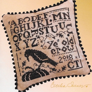 May include: A beige cushion with a black embroidered alphabet, a black crow, and the text "Ye old crow" and "2018 CT". The cushion has a black pom-pom trim.