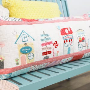 May include: A rectangular cushion with an embroidered candy shop scene. The white cushion features colourful embroidered shops, lollipops, and a heart-shaped balloon. It has a red and white striped border and is placed on a blue bench.