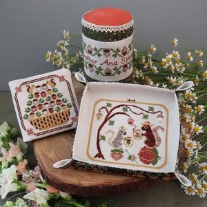 May include: A set of three needlepoint pieces featuring a squirrel theme. The pieces include a pincushion, a small square needlepoint, and a larger square needlepoint with a basket of acorns. The pincushion is red and white and has the text "Gather And Stitch" embroidered on it. The small square needlepoint features a basket of acorns with squirrels. The larger square needlepoint features two squirrels sitting under a tree with acorns. The pieces are displayed on a wooden surface with flowers.