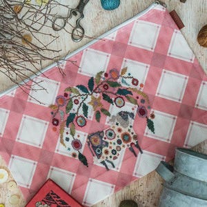 May include: Pink and white checkered zippered pouch featuring a cross-stitched sheep design with floral accents. The sheep is adorned with colorful patterns. The pouch is rectangular and has a white zipper.