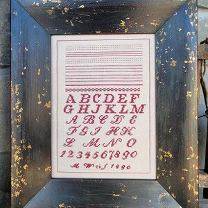May include: A framed cross-stitch sampler with a black frame with gold accents. The sampler features a red alphabet, numbers, and the text "M.W.c.f. 1890".