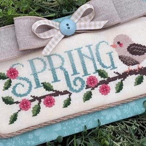 May include: Cross-stitched rectangular artwork with the word "SPRING" in teal, a bird, and floral branch. The artwork is framed with a rope-like border and topped with a beige bow and a light blue button. The background is a floral pattern.