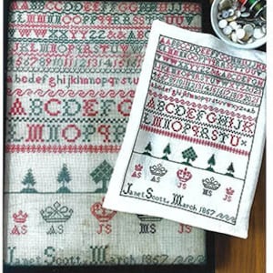 May include: A white cross-stitch sampler with the text "Janet Scott March 1857" and various letters of the alphabet, crowns, and trees. The sampler is framed and displayed on a wooden surface.