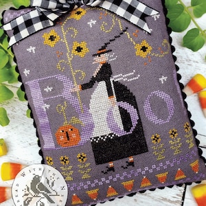 May include: A purple cross-stitch pillow with a black and white checkered ribbon. The pillow features a witch holding a broom and a pumpkin with a face. The text "BOO" is stitched in purple on the pillow. The pillow has a black scalloped border.