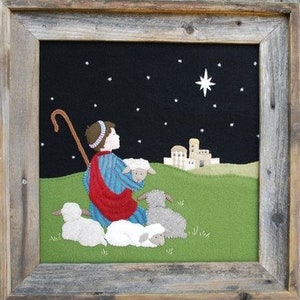 May include: A felt Christmas scene depicting a shepherd kneeling with his sheep, looking up at a bright star in the night sky. The scene is framed in a rustic wooden frame.