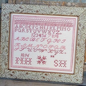 May include: A framed cross-stitch sampler with a white background and red stitching. The sampler includes the alphabet, numbers, and the year 1896. The sampler is framed in a white, ornate frame.