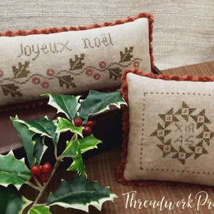 May include: Two embroidered pillow covers with a Christmas theme. The first pillow cover is beige with a red fringe and says "Joyeux Noel" in French with a green vine and red berries. The second pillow cover is beige with a red fringe and has a green starburst design with the letters "XMAS" in the center.