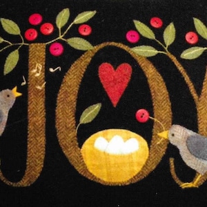 May include: A felt wall hanging with the word "JOY" in brown letters, decorated with two birds, a heart, and a nest with eggs. The background is black and the branches are green with red berries.