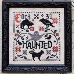 May include: A framed cross-stitch with a Halloween theme. The design features a black cat, raven, ghost, and the word "HAUNTED." The date "Oct 31" is displayed with floral and star accents, in black, orange, and grey.