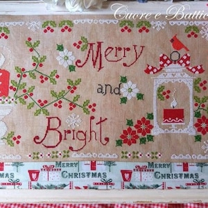 May include: Cross stitch pattern featuring a lantern with a red candle, holly, and the text "Merry and Bright". The pattern is framed and has a red and white gingham border.