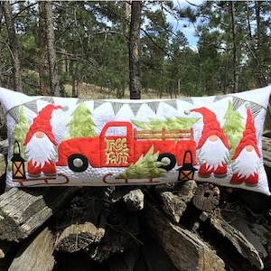 May include: A red truck with a Christmas tree in the back, two gnomes on either side, and the words "Tree Farm" on the side. The truck is on a white pillow with a green and white garland.