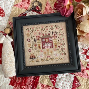 May include: A framed cross-stitch embroidery depicting a red house with a garden. The embroidery is on a beige linen fabric with a black frame. The text "GARDEN" is stitched in the upper left corner of the embroidery.