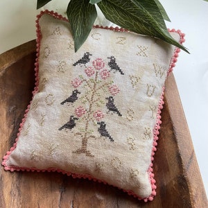 May include: A cream-colored pillow with a pink pom-pom trim features an embroidered design of a tree with black birds and pink flowers. The design includes the letters of the alphabet.