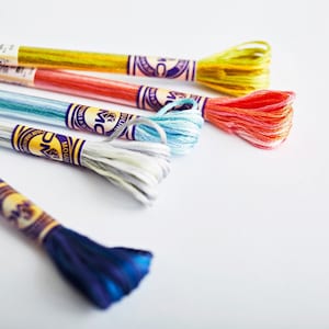 May include: Six spools of embroidery floss in various colors, including yellow, orange, blue, and white. The spools are labeled with the brand name "DMC Mouliné" and the color number.