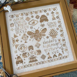 May include: A framed cross-stitch embroidery featuring a bee, a beehive, flowers, and two sets of the alphabet in brown thread on a white background.