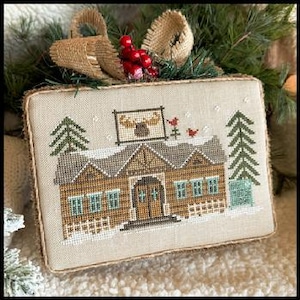 May include: A rectangular cross-stitch wall hanging on beige fabric featuring a brown cabin with a moose sign, two green pine trees, and red birds. The piece is framed with a burlap border and topped with a burlap bow and red berries.