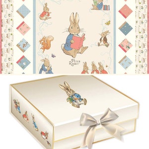 May include: A cream-coloured gift box adorned with Peter Rabbit illustrations. The box has a gold trim and a satin bow. The lid features a Peter Rabbit image, and the sides display other character illustrations.