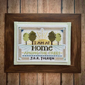 May include: A framed cross-stitch with a white background and green, brown, and yellow embroidery. The embroidery depicts three trees with a sun rising behind them. The text reads "I am at home among the trees" and "J.R.R. Tolkien".
