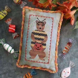 May include: A cross-stitch pillow featuring a brown bear dressed as a bee holding a jack-o-lantern. The pillow is surrounded by colorful candy.