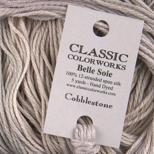 May include: Close-up of a skein of Classic Colorworks Belle Soie thread in a light gray color, labeled "Cobblestone." The label indicates it is 100% 12-stranded spun silk, 5 yards, and hand-dyed.