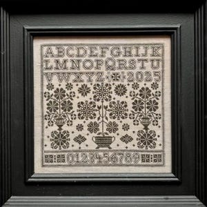 May include: Framed cross-stitch sampler with a black frame. The sampler features an alphabet, numbers, and floral motifs in shades of brown and beige. The year 2025 is stitched into the design.