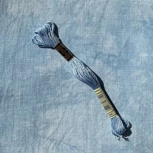 May include: A skein of light blue embroidery floss rests on a matching blue fabric background. The floss is wrapped with a brown paper label and a barcode. The fabric has a textured appearance.