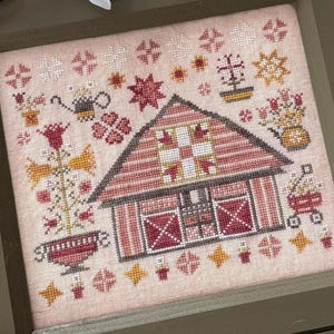 May include: A pink and white cross-stitch embroidery depicting a red barn with a patchwork quilt on the roof. The barn is surrounded by flowers, a watering can, and other small details.