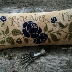 May include: A beige pillow with a cross-stitch design featuring a floral motif with blue roses, green leaves, and two birds. The text "Remember Me" is stitched in black thread. The pillow is labeled "Scattered Seed Samplers C2020".