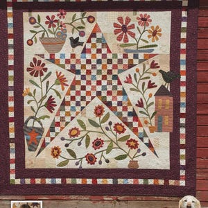 May include: A handcrafted quilt with a central star design, featuring floral and bird appliqués. The quilt showcases a patchwork star in the center, surrounded by a cream-colored background and a burgundy border. The quilt is displayed against a red wooden backdrop.