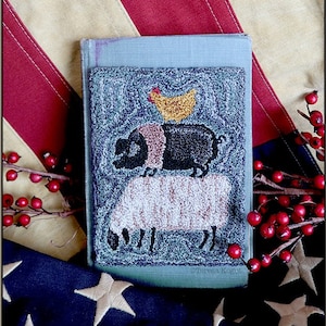 May include: A hand-stitched needlepoint piece depicting a pig, a sheep, and a chicken. The piece is framed in a blue book cover and set against a red, white, and blue American flag.