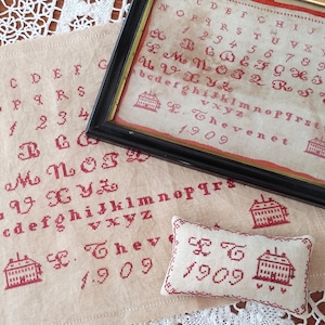 May include: Two framed pieces of needlepoint embroidery with red lettering and a small house design. The larger piece includes the alphabet, numbers, and the text "L. Thevenet 1909". The smaller piece includes the text "L.T. 1909" and a small house design.