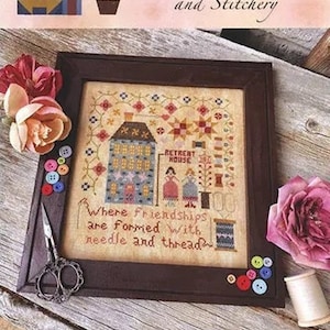 Peut inclure: Un motif de point de croix encadré représentant une maison avec le texte "Retreat House" et la phrase "Where friendships are formed with needle and thread". Le titre "Stitcher's Retreat House" est situé sous le cadre, avec le nom du créateur "Lori Pengelly".