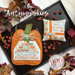 May include: A cross-stitch embroidery design featuring a pumpkin with the text "Autumn Skies & Pumpkin Pies" and "This Way to the Pumpkin Patch". The design is framed with a brown felt border and includes a small, decorative bow.