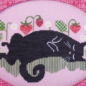 Peut inclure: Broderie au point de croix représentant un chat noir se prélassant au milieu de fraises et de fleurs. Le chat a des moustaches et des détails blancs, sur fond rose. Le motif est encadré d'une bordure rose à motifs.