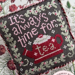 May include: A gray and red cross-stitch pillow with a teapot and the text "It's always tea time,  "