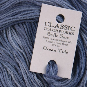 May include: Close-up of a skein of blue yarn with a tag. The tag reads "CLASSIC COLORWORKS Belle Soie" and "Ocean Tide." The yarn is 100% 12-stranded spun silk, 5 yards, and hand dyed. The yarn is a soft, muted blue.