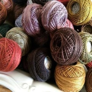 May include: A collection of colorful thread spools in various shades of red, brown, green, and gold. The spools are tightly wound and appear to be made of a fine, textured material, suitable for embroidery or other needlework projects.