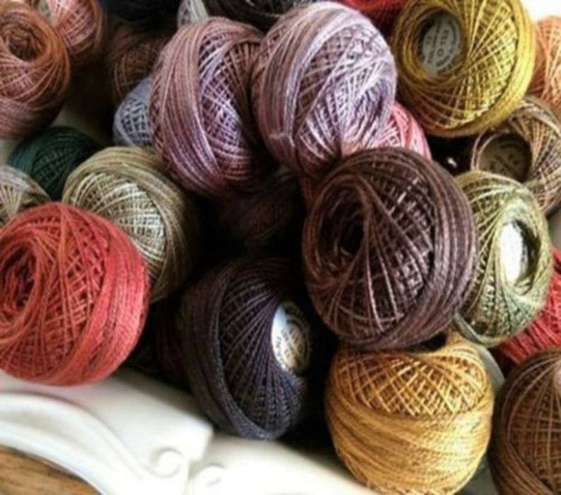 May include: A collection of colorful thread spools in various shades of red, brown, green, and gold. The spools are tightly wound and appear to be made of a fine, textured material, suitable for embroidery or other needlework projects.